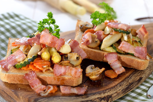 Deftiger Toast mit gebratenem Gemüse und Speck, auf einem Holzbrett serviert - Hearty toast with fried vegetables and bacon, served on a wooden board