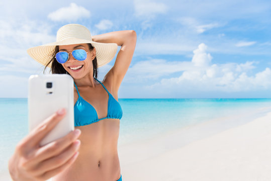 Summer beach vacation girl taking fun mobile selfie photo with smartphone. Cute Asian woman wearing blue sunglasses posing for phone self-portrait photo enjoying suntan in tropical travel holidays.