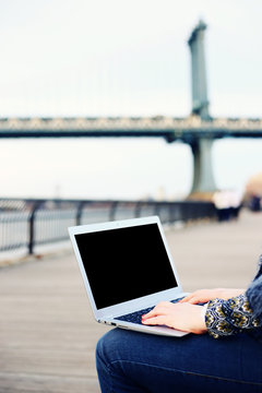 Young woman  sitting using a laptop. Black screen. Copy space. High key, bright crisp high resolution image. Brooklyn Heights promenade. Manhattan bridge, New York. 