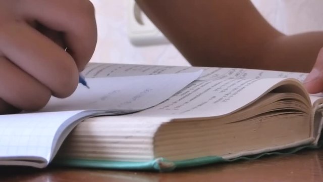 On the table lies a thick textbook, a hand with a pen in a notebook record homework