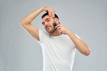 Naklejka premium smiling man shaving beard with trimmer over gray