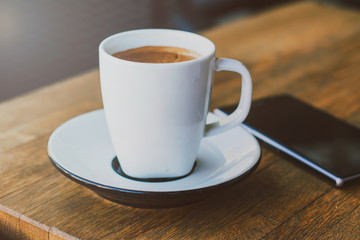A cup of coffee and smart phone on a woden table in cafe, soft warm filter, lens flare