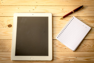 Tablet, notebook and pen resting on a wooden table with soft lig
