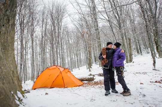 Happy climbers couple standing next to their winter mountains base camp in the forest, man and woman are looking at each other. Climbing equipment.