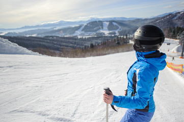 Side portrait of young cute woman skier looking at beautiful mountain landscape at the ski resort on a sunny day. Girl is wearing blue jacket helmet and goggles. Carpathian Mountains, Bukovel