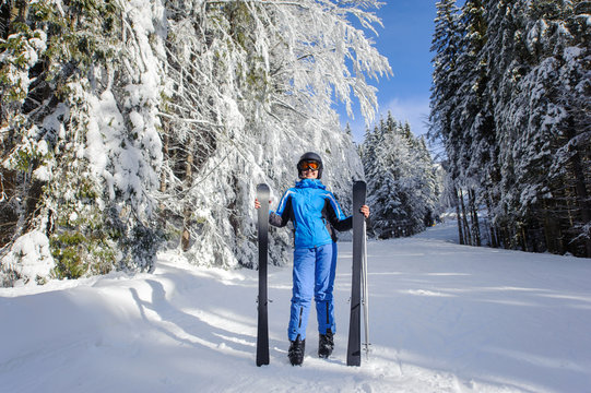 Full length portrait of smiling happy young female skier in winter forest. Woman is wearing blue ski suit helmet and orange glasses, holding skis. Winter sports concept. Carpathian Mountains, Bukovel