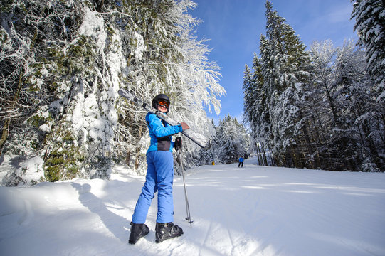 Full length portrait of young happy female skier on a ski slope in the winter forest on a sunny day. Woman is holding skis on her shoulder smiling and looking towards the camera. Ski resort. Bukovel
