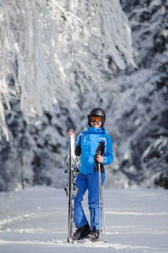 Portrait of young happy female skier standing enjoying sunny day against beautiful snow covered trees on the background. Woman is holding her skis in one hand and ski poles in another. Bukovel
