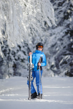 Isolated portrait of young happy female skier standing enjoying sunny day against beautiful snow covered trees on the background. Girl is holding her skis in one hand and poles in another. Bukovel