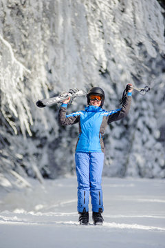 Full length portrait of young happy female skier standing enjoying sunny day against beautiful snow covered trees on the background. Girl is holding her skis and ski poles up. Bukovel, Ukraine