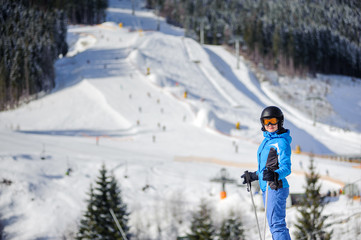 Young happy woman skier against ski slopes and ski-lift on background. Woman is wearing helmet skiing glasses gloves and blue ski suit. Winter sports concept.