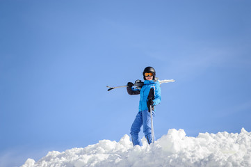 Portrait of young female skier standing on top of the mountain against blue sky on a sunny day. Woman is holding skis on her shoulder smiling and looking into the camera. Winter sports concept.