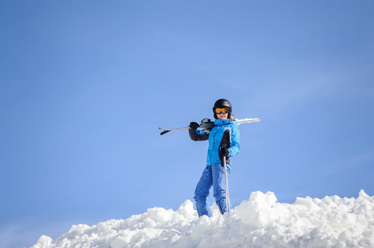 Portrait of young female skier standing on top of the mountain against blue sky on a sunny day. Woman is holding skis on her shoulder smiling and looking into the camera. Winter sports concept.