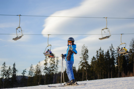 Happy young female skier on a sunny day at ski resort against ski-lift. Girl is standing on a ski slope and giving the thumb up. Winter vacation.