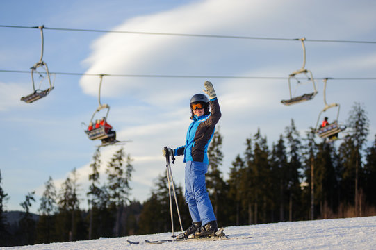 Happy young female skier on a sunny day at ski resort against ski-lift. Girl is standing on a ski slope with raised arms as sign of success. Winter vacation. Carpathian Mountains, Bukovel