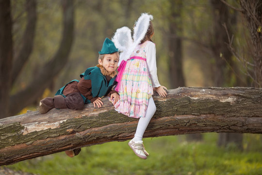 Portrait of a cute little boy in knight costume sitting on a tree in a park with a girl dressed up as a fairy