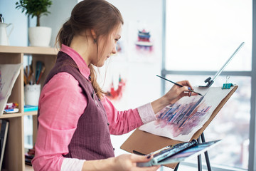Side view portrait of a young woman painter drawing cityscape with watercolor palette on white...