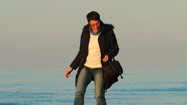 Woman in blue jeans and dark brown coat walking on the beach and picking pebbles
