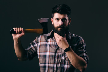 Young bearded man with big axe and looking at camera while standing against dark background