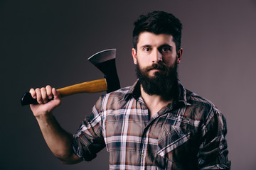 Young bearded man with big axe and looking at camera while standing against dark background