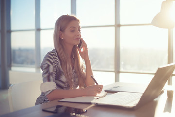 Side view portrait of young businesswoman having business call in office, her workplace, writing...