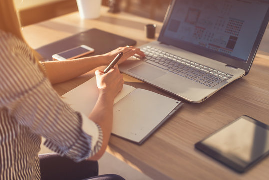 Female designer using laptop, sketching at blank notepad. Woman hand writing in notebook on wooden desk. - Powered by Adobe