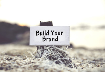 concept image, word BUILD YOUR BRAND on white canvas frame with blurred beach and clam shell background.