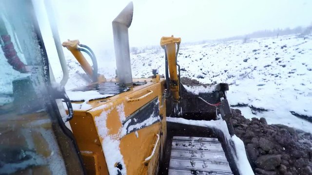 Bulldozer machine working with ground on snow winter day. View from cabin