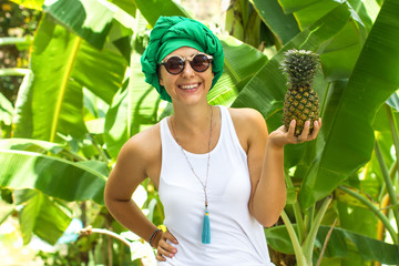 The young girl, holds Pineapple in hand and smiles, Dressed in bright clothes and with the rolled-up scarf on the head, the fashionable woman, in sunglasses