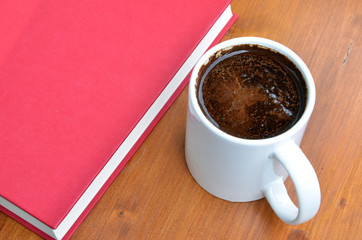 White cup of coffee and red book on brown wooden desk in sunshine