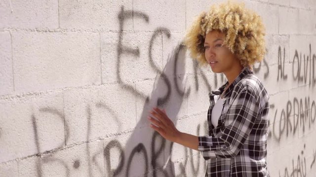 Young mixed-race woman dressed in plaid men's shirt and white top walking along a graffiti sprayed stone wall and looks later back in slow motion. 