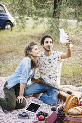 Young couple in love doing a picnic outdoors in Tuscany wine country and taking a selfie with an instant camera