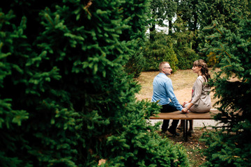 Beautiful pregnant stylish couple relaxing outside in the autumn park sitting on bench. They are lovely looking at belly. Copy space