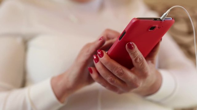 Woman looks and flips through the photos in her smartphone. Female with smartphone. Close up of woman hands with red smartphone. Businesswoman with red mobile phone with earphones