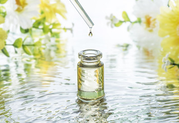 The bottle of Moisturizing cosmetic oil in the water waves on the summer flowers blur background and  pipette with oil drop above the bottle - obrazy, fototapety, plakaty
