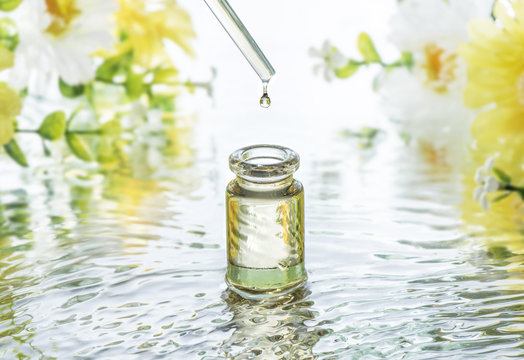 The Bottle Of Moisturizing Cosmetic Oil In The Water Waves On The Summer Flowers Blur Background And  Pipette With Oil Drop Above The Bottle