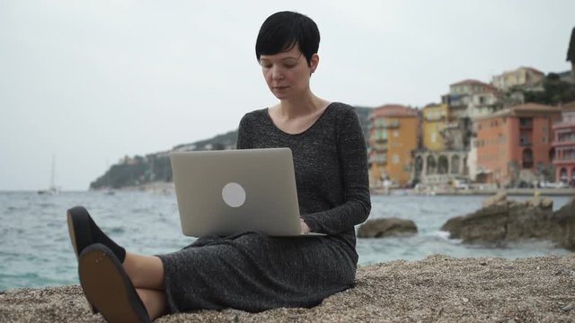 Frenchwoman using laptop at her vocation in Nice Cote d'Azur Provence. Female sitting on the beach read news check mail. On the backgroung view city.