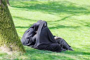Group of muslim women in hijab on green grass in city park, looking film on smatrfone.