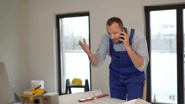 Angry male worker with blueprints talking on cellphone at new home
