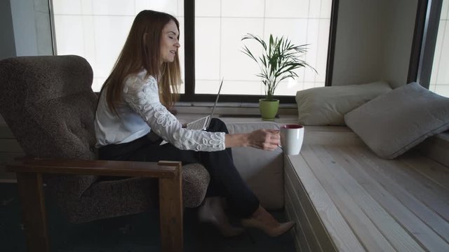Female talking and gesturing. At the end conversation she waving goodbye. She dressed formal white blouse. On her friendly face happy smile. She holding cup in hands with nude nail polish.