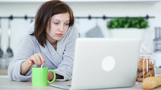 Sleepy woman stirs a coffee in the cup and periodically fall asleep. Use the laptop at the kitchen