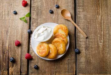 Cottage cheese pancakes with sour cream, raspberries, blueberries and mint, homemade traditional Ukrainian and Russian syrniki on rustic wooden background. Top view.