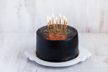 Black round birthday cake with burning golden candles on white b