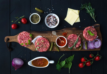 Ingredients for cooking burgers. Raw ground beef meat cutlets on wooden chopping board, red onion, cherry tomatoes, greens, pickles, tomato sauce, cheese, herbs and spices over black background, top