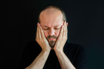 Portrait of an adult man with beard and mustaches on black background, eyes closed, having headache