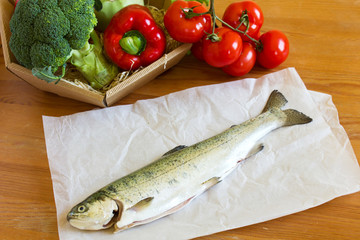Raw fresh trout lying on a wooden table with a basket of vegetab