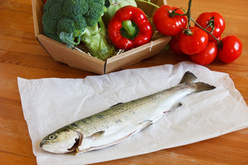 Raw fresh trout lying on a wooden table with a basket of vegetab