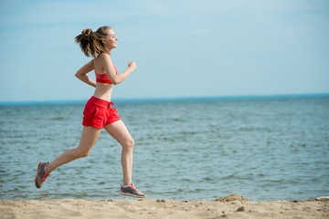 Fototapeta premium Young lady running. Woman runner running at the sunny summer sand beach. Workout near ocean sea coast. Beautiful fit girl. Fitness model caucasian ethnicity outdoors. Weight loss exercise. Jogging.