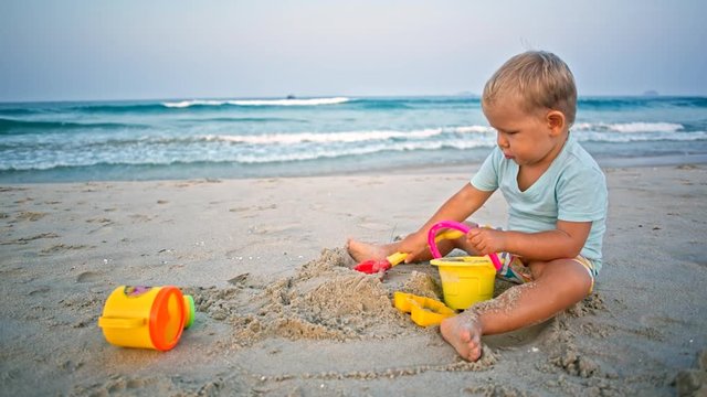 toddler boy plays sand toys on the beach with sea tide in background wide angle shot