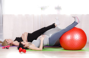 Two happy pregnant women,brunette and blonde,dressed in sportswear,execute a complex of exercises for pregnant women with large gymnastic balls,grey and red,  fitness centre